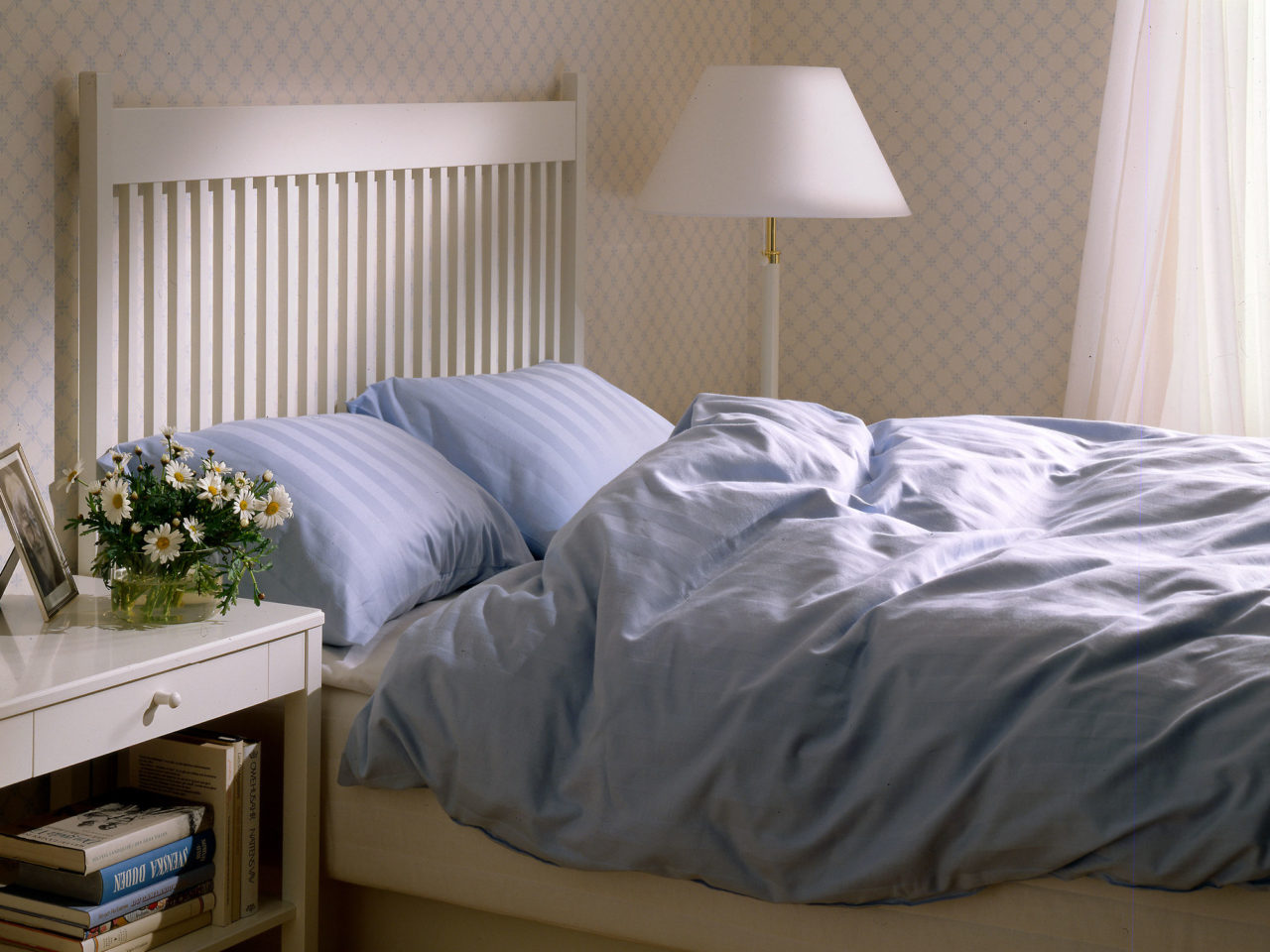 Ett sovrum med möbler och detaljer i vita nyanser. Dubbelsängen, modell STOCKHOLM, har hög gavel och ljusblå sängkläder.