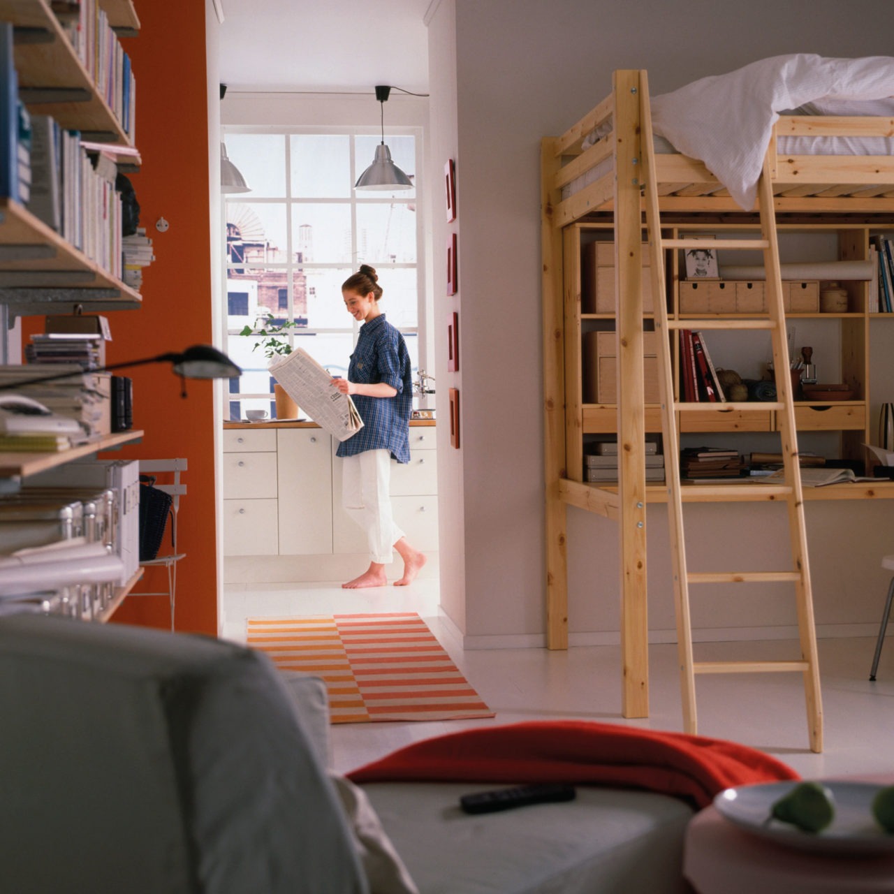 Habitación con cama elevada y espacio de trabajo, estanterías sujetas a la pared, diván y paso hacia la cocina.