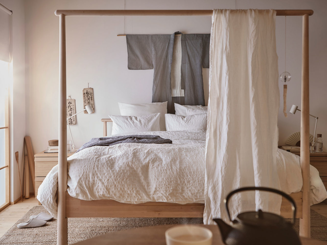 Schlafzimmer mit Doppelbett aus hellem Holz. Fußende mit hohem, überdimensioniertem Rahmen und leichtem, drapiertem Stoff.