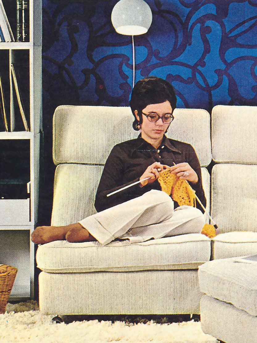 Sur fond de mur à motifs bleus, une femme tricote sur un canapé deux places blanc à haut dossier, encadré de bibliothèques.