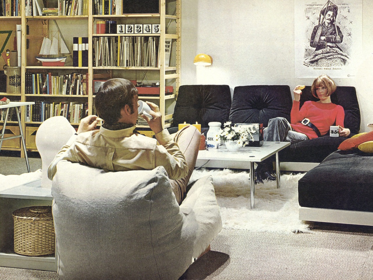 Ett vardagsrum där en man i en kuddfåtölj och en kvinna i en svart soffa dricker kaffe på varsin sida om ett lågt soffbord.