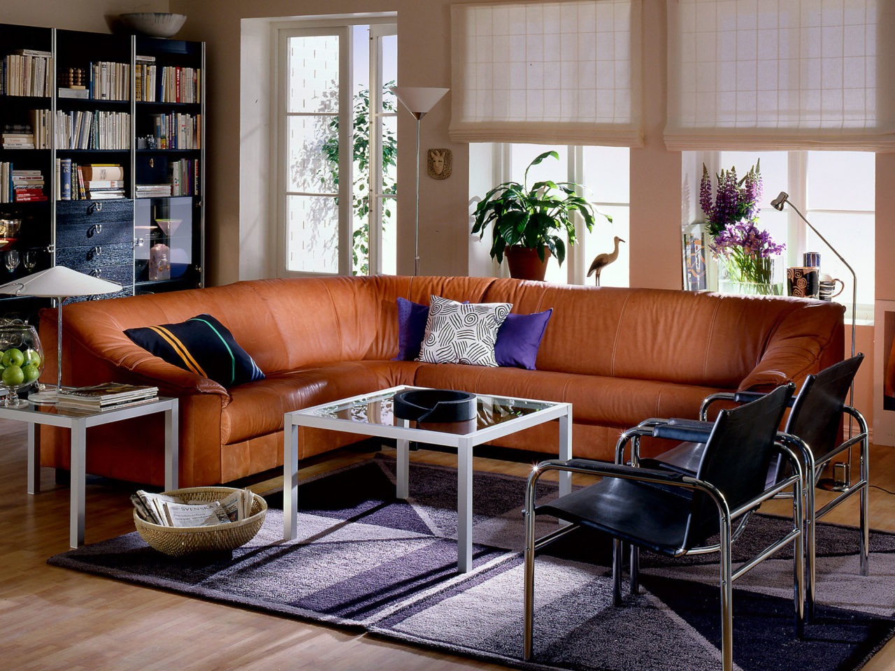 Salon parqueté avec grand canapé d’angle en cuir marron. À côté, deux chaises chrome et cuir noir.