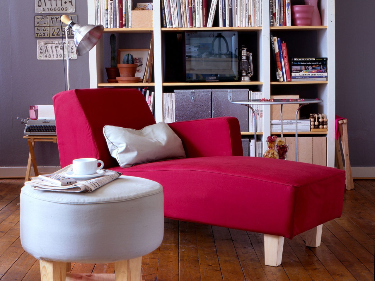 Cerca de una estantería hay un diván rojo robusto y sencillo. Al lado, un taburete redondo y claro de diseño similar.