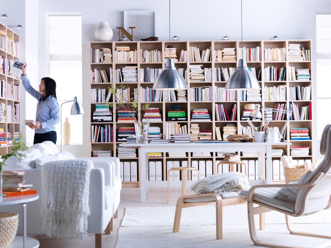 Helles Wohnzimmer mit Bücherregalen über die gesamte Wandlänge. Mehrere Sessel, darunter POÄNG mit Fußhocker.