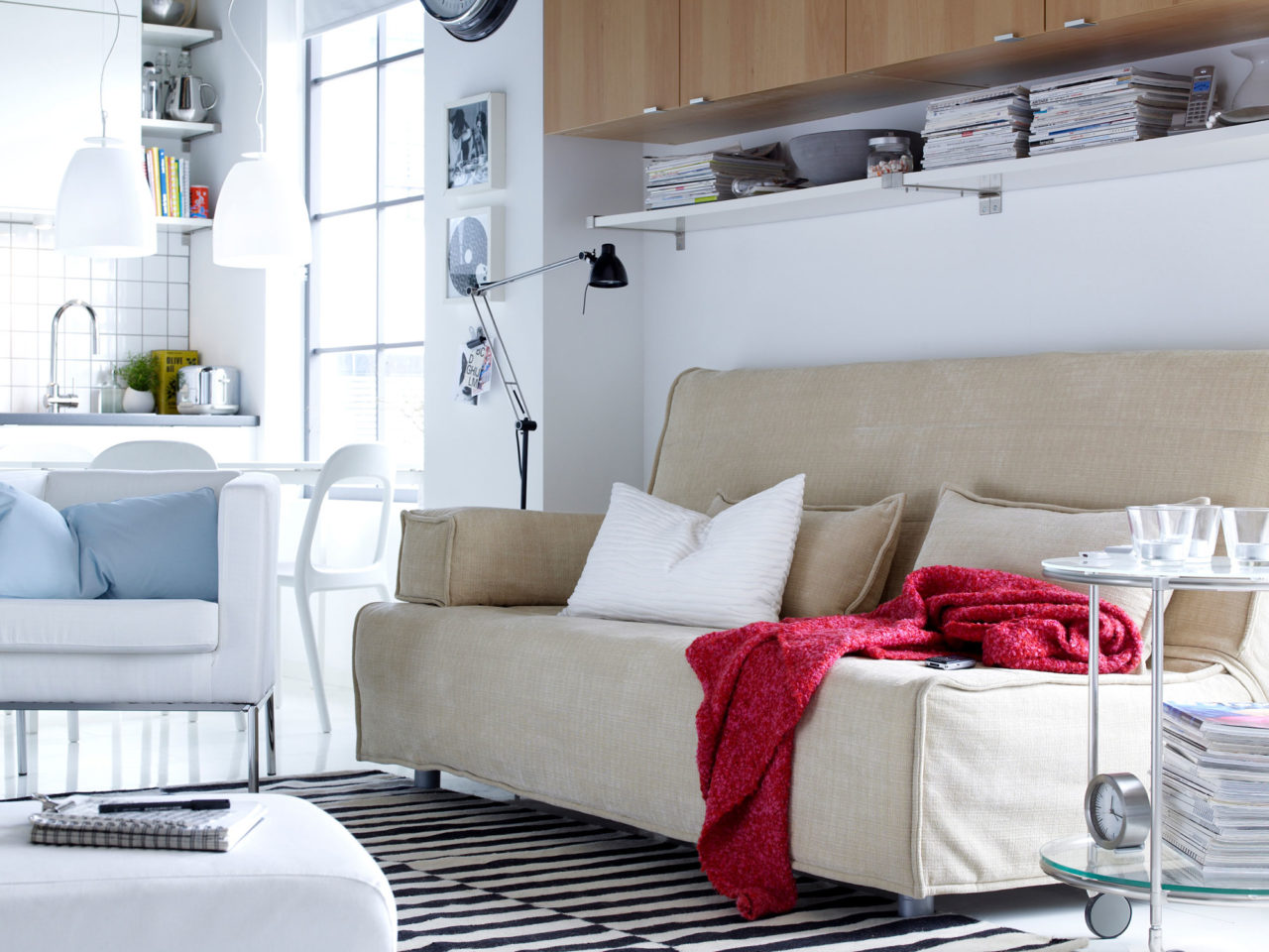 Canapé et fauteuil clairs sur un tapis à rayures noires et blanches. Le salon est placé à côté d’une cuisine toute blanche.