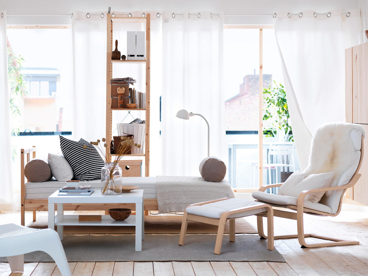 Ett ljust rum med solbelysta golv till tak-fönster, en säng och hylla i ljust trä samt en fåtölj och fotpall, modell POÄNG.