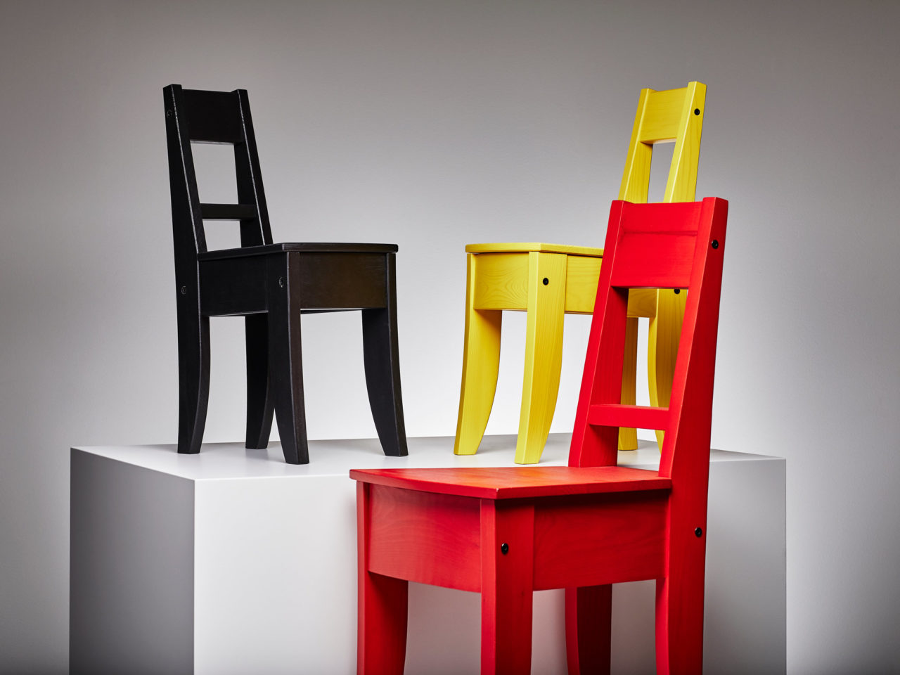 Three simple wooden chairs, one black, one yellow, one red, arranged on and next to a white cube.