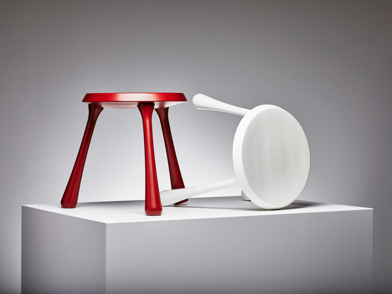 Two three-legged stools, one red, one white, on top of a white cube.