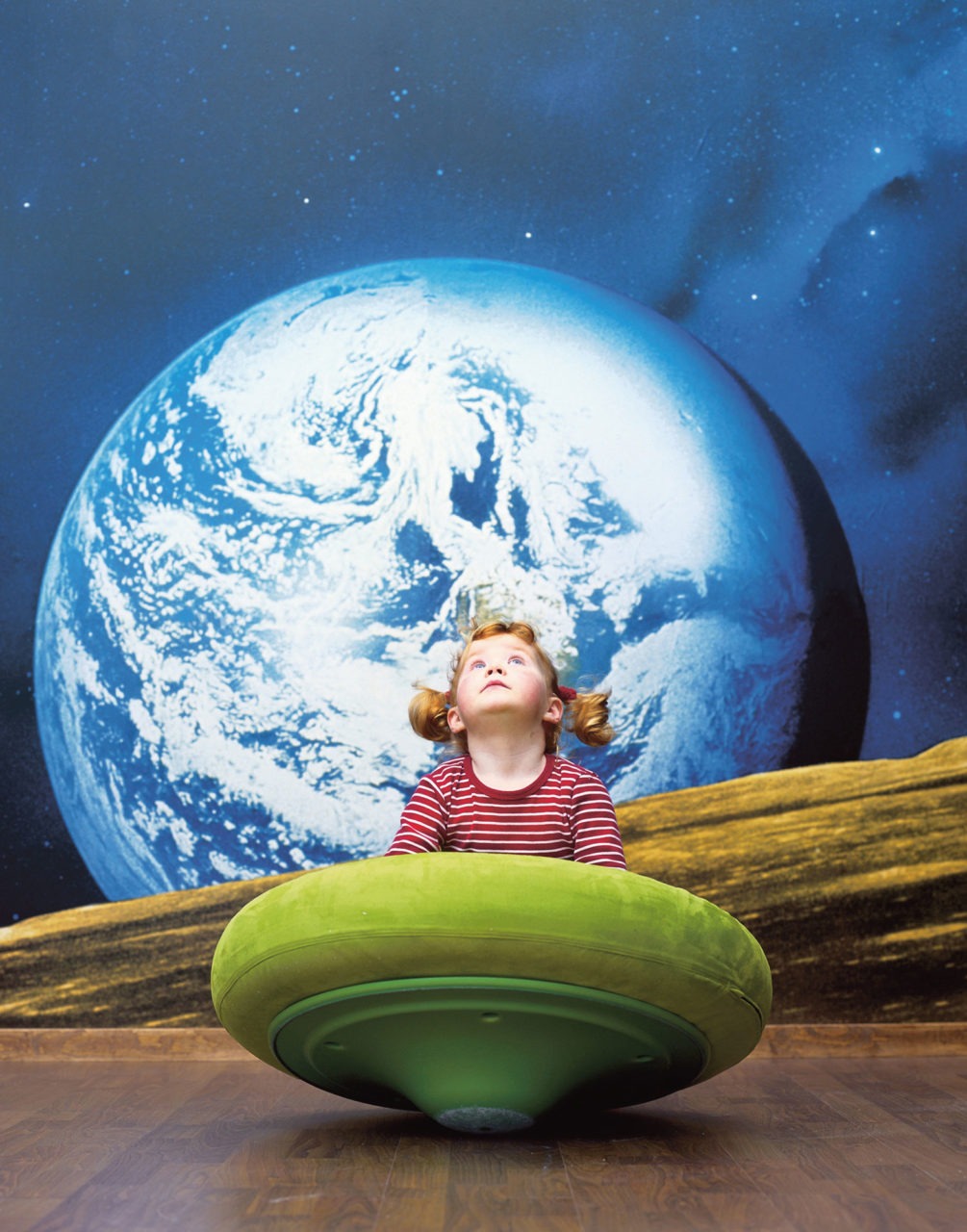 Liten flicka leker på grön planetformad möbel, i bakgrunden syns stor bild av jorden sedd från rymden.