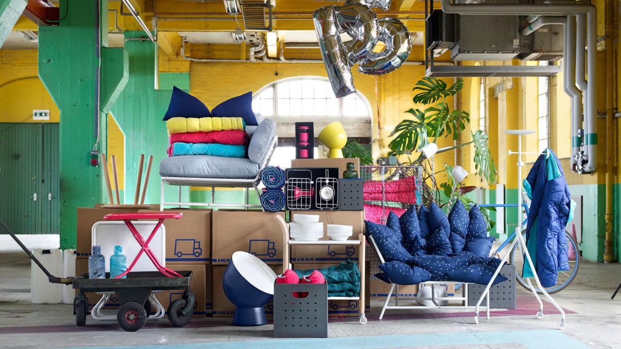 Möbler, mattor och kuddar i olika färger staplade på varandra i lagerlokal, två silverfärgade ballonger bildar ordet PS.