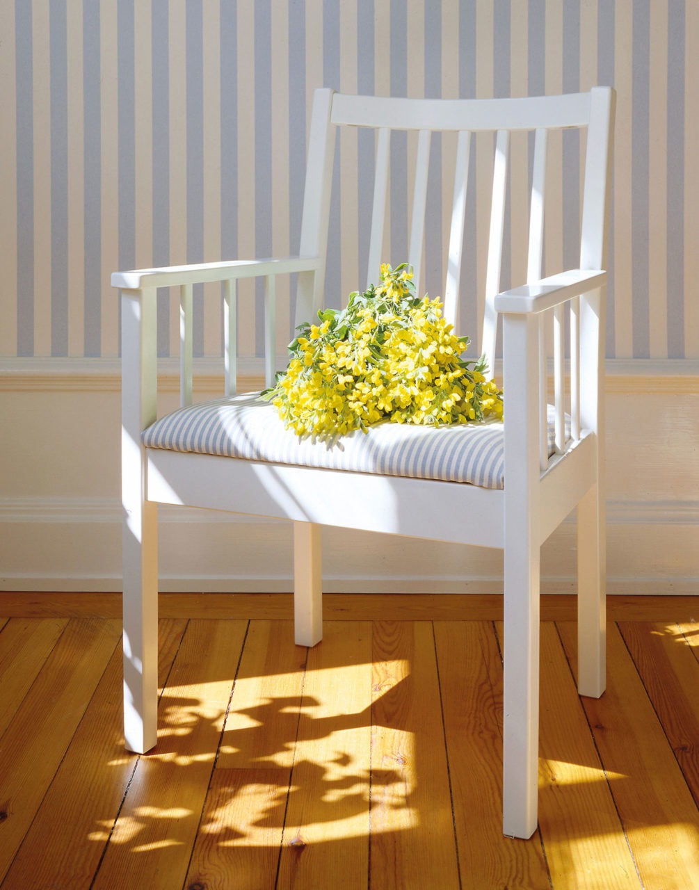White wooden armchair stands on harwood floor, a bouquet of yellow flowers is placed on the blue-white-striped seat cushion.