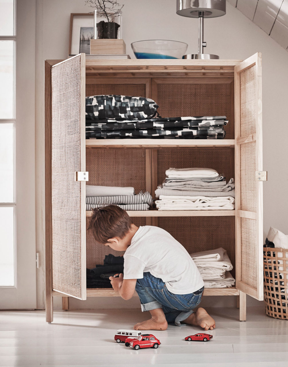 Small boy squatting by open linen cupboard made of light wood. On the floor are some small red toy cars.