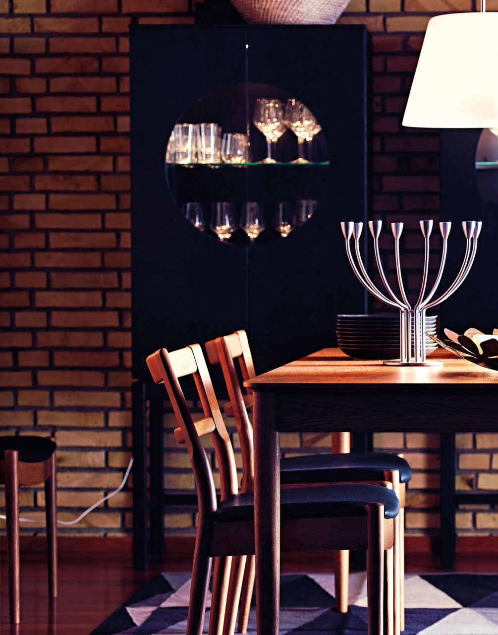 Dining room with brick wall, wooden table and chairs, black display cabinet with wine glasses behind round windows.