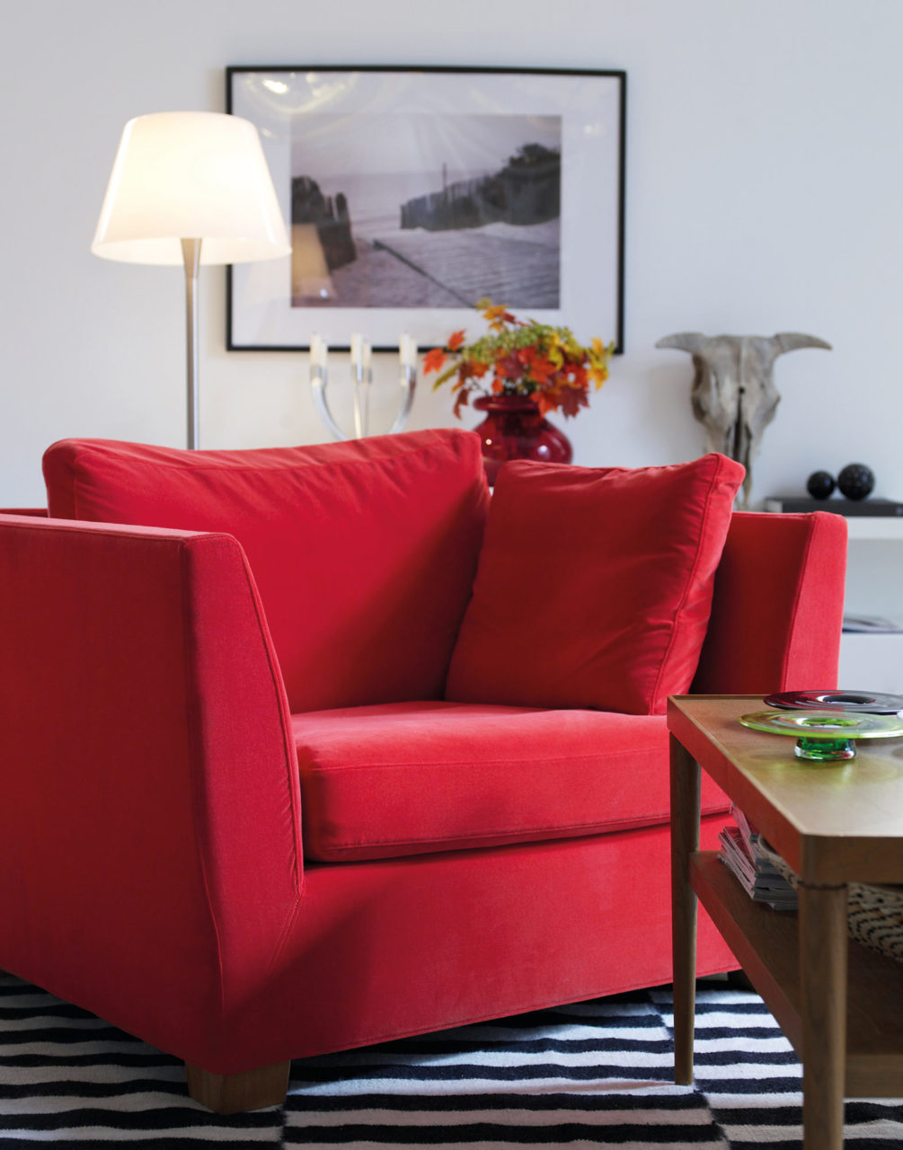 Stor röd sammetsfåtölj och brunt soffbord, i bakgrunden en tänd golvlampa och tavla på väggen.