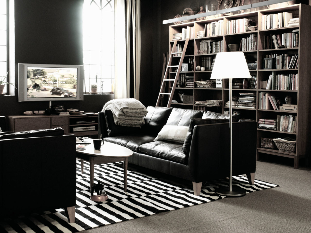Vardagsrum med mörkgröna väggar, två svarta soffor och ovalt soffbord. Hög bokhylla täcker ena väggen.