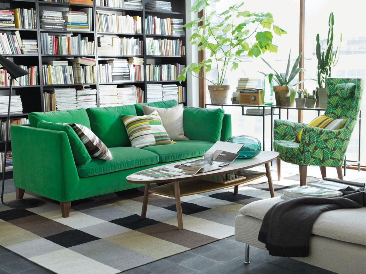 Vardagsrum med gröna växter i stora fönster, soffa och fåtölj i gröna toner, bokhyllor täcker en hel vägg.