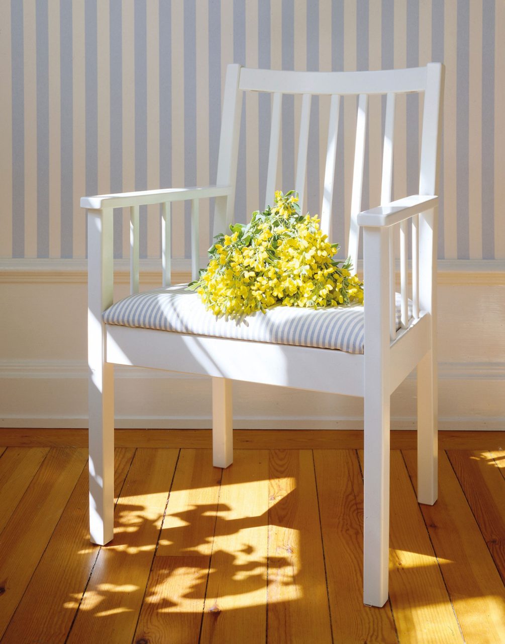 Vit karmstol i trä står på trägolv, en bukett med gula blommor ligger på den blåvitrandiga sittdynan.
