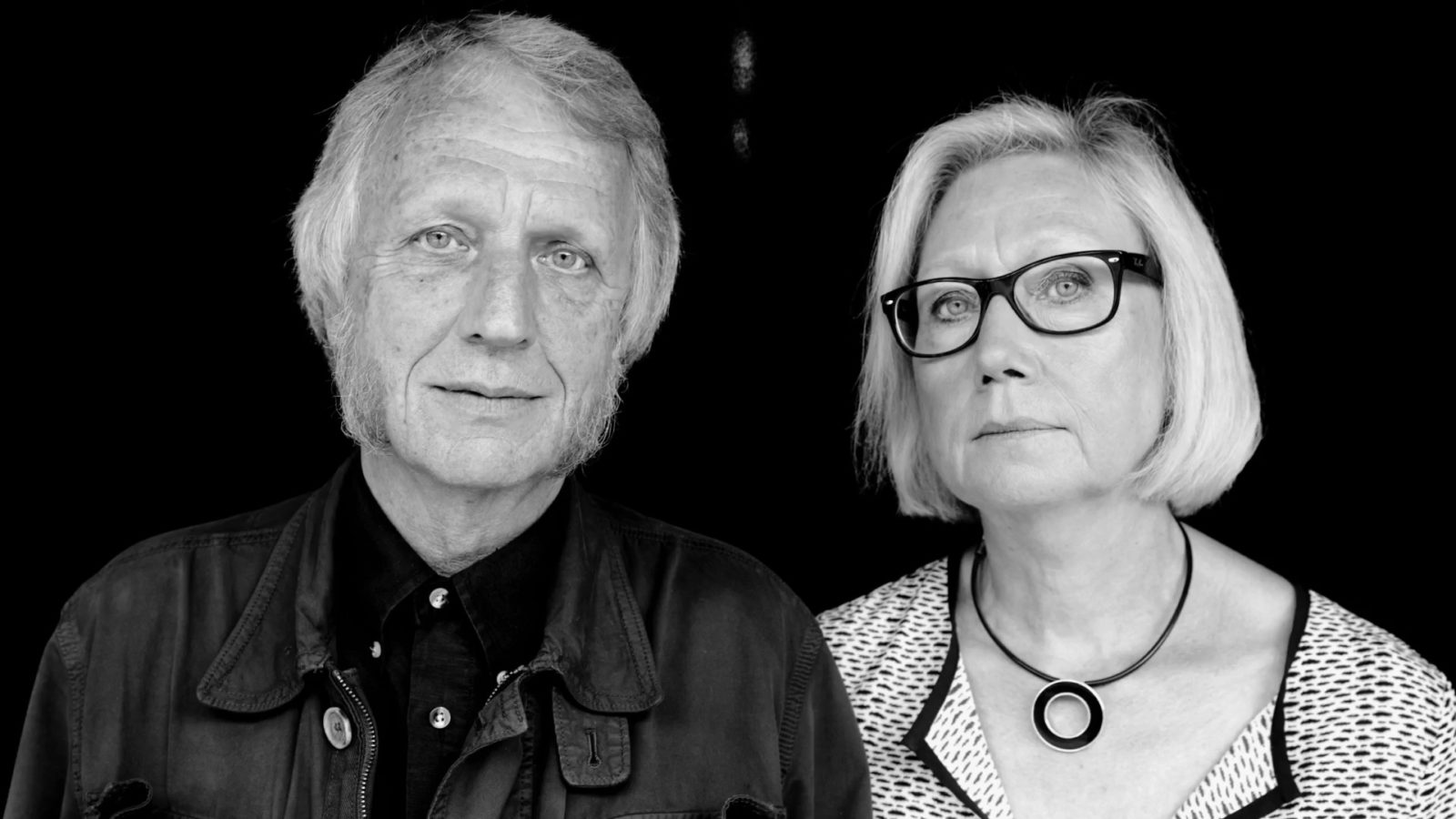 Ageing man and woman dressed in black and white, Knut Hagberg and Marianne Hagberg, carrying simple wooden table together.