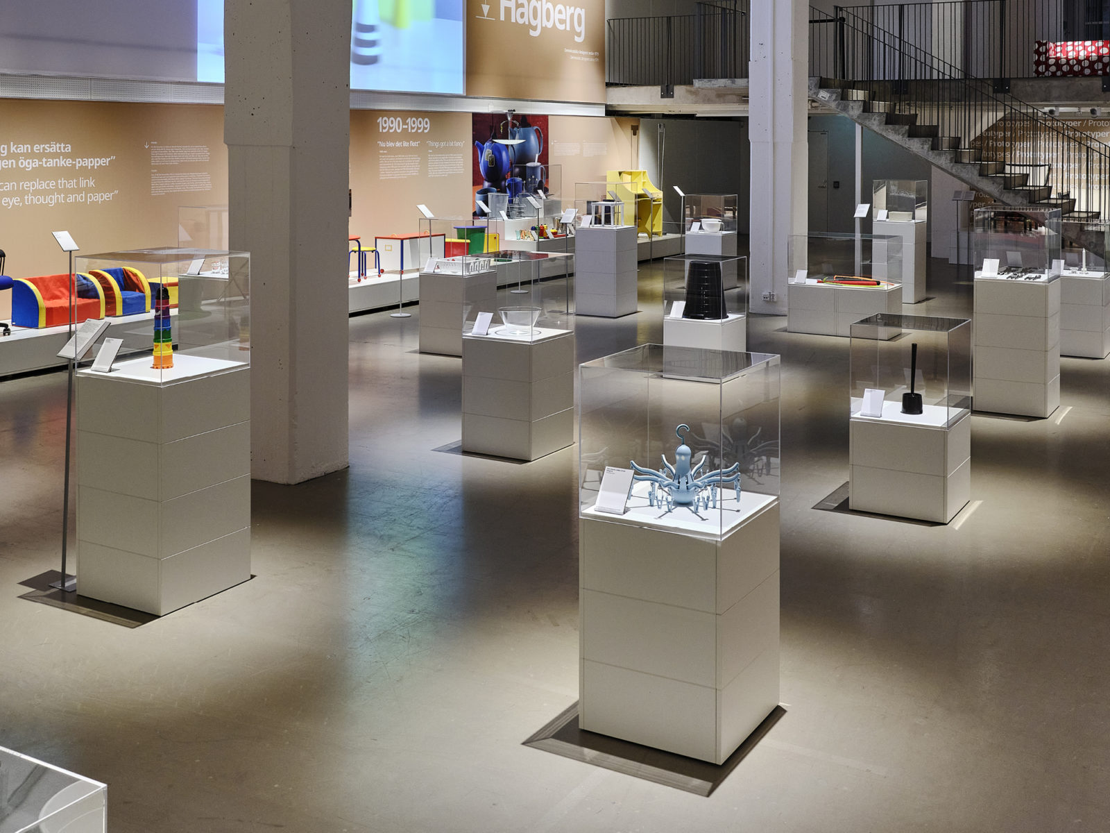 Exhibition hall where objects are displayed in glass cabinets on white cubes, and images are projected onto walls.