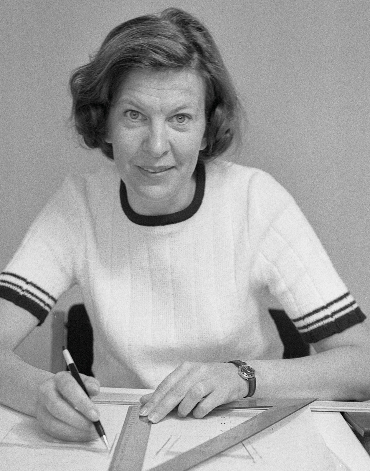 Karin Mobring, dark haired in white knitted T-shirt sits at drawing table working with pen and ruler.