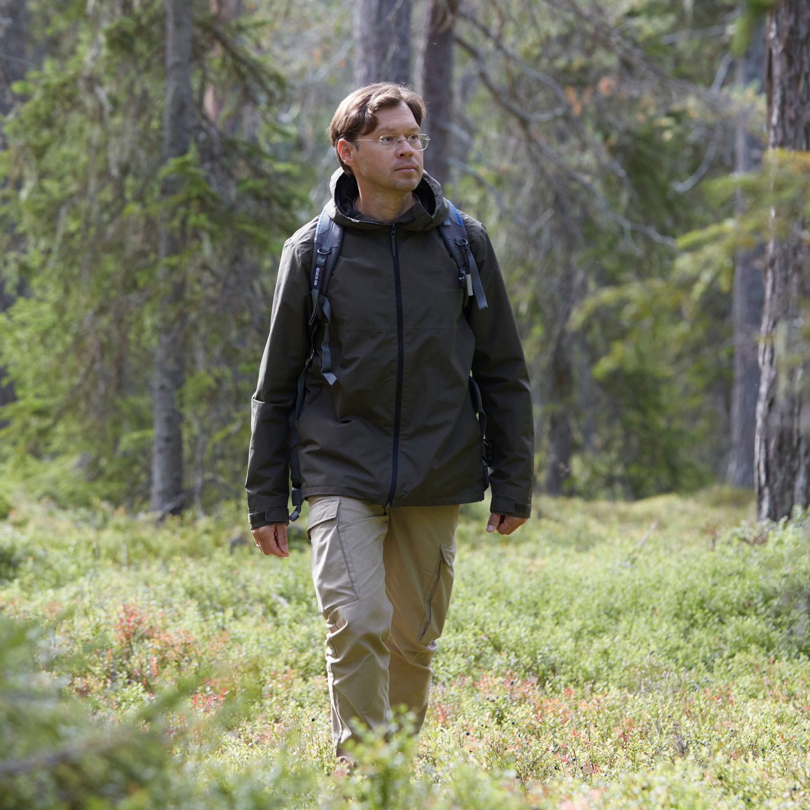 Mörkhårig man, Mikhail Tarasov, med stålbågade glasögon och vädertåliga kläder går i skogen.