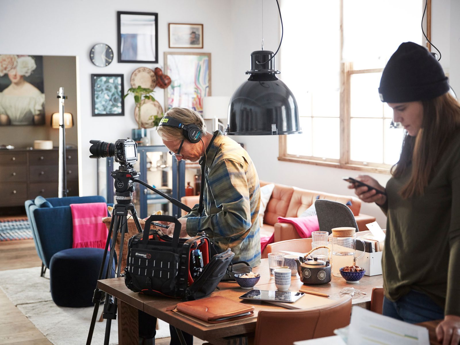 Produktionsteam, man och kvinna, står med en kamera på stativ runt ett bord i ljust rum med modern inredning.
