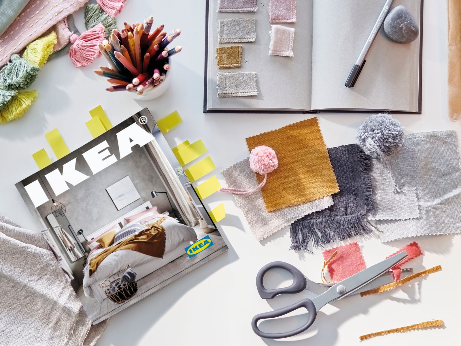 Bordsskiva sedd uppifrån med IKEA katalog med postitlappar, skissblock, tygprover, färgpennor och sax.
