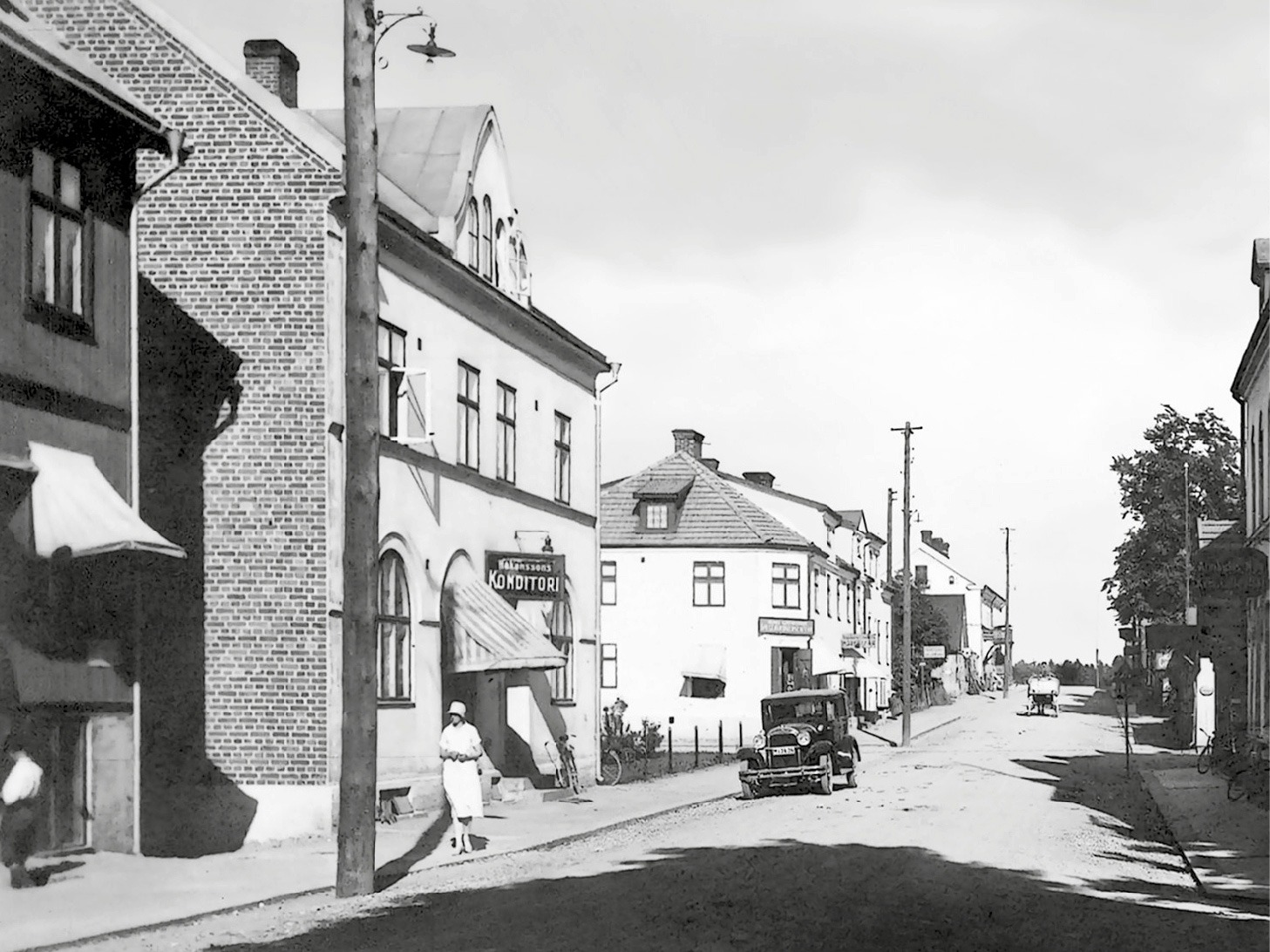 Quiet Swedish street in the 1930s, a woman dressed in white on the sidewalk, one parked car, one car coming down the street.
