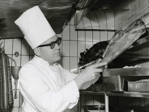 Ulf Renström, con gafas y ropa de cocinero, ante una pierna de cordero en la cocina de un restaurante.