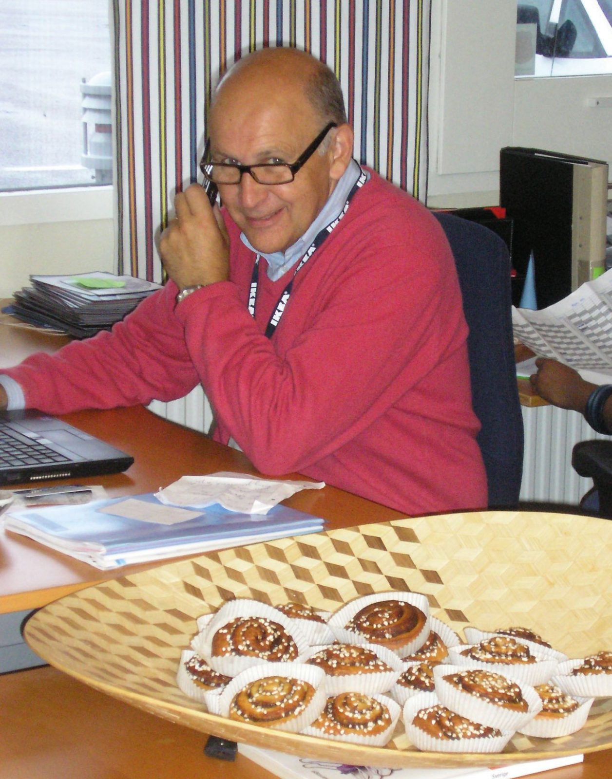 Man wearing glasses and red sweater, Mats Agmén, is on the phone at a desk, in foreground a big basket with cinnamon buns.