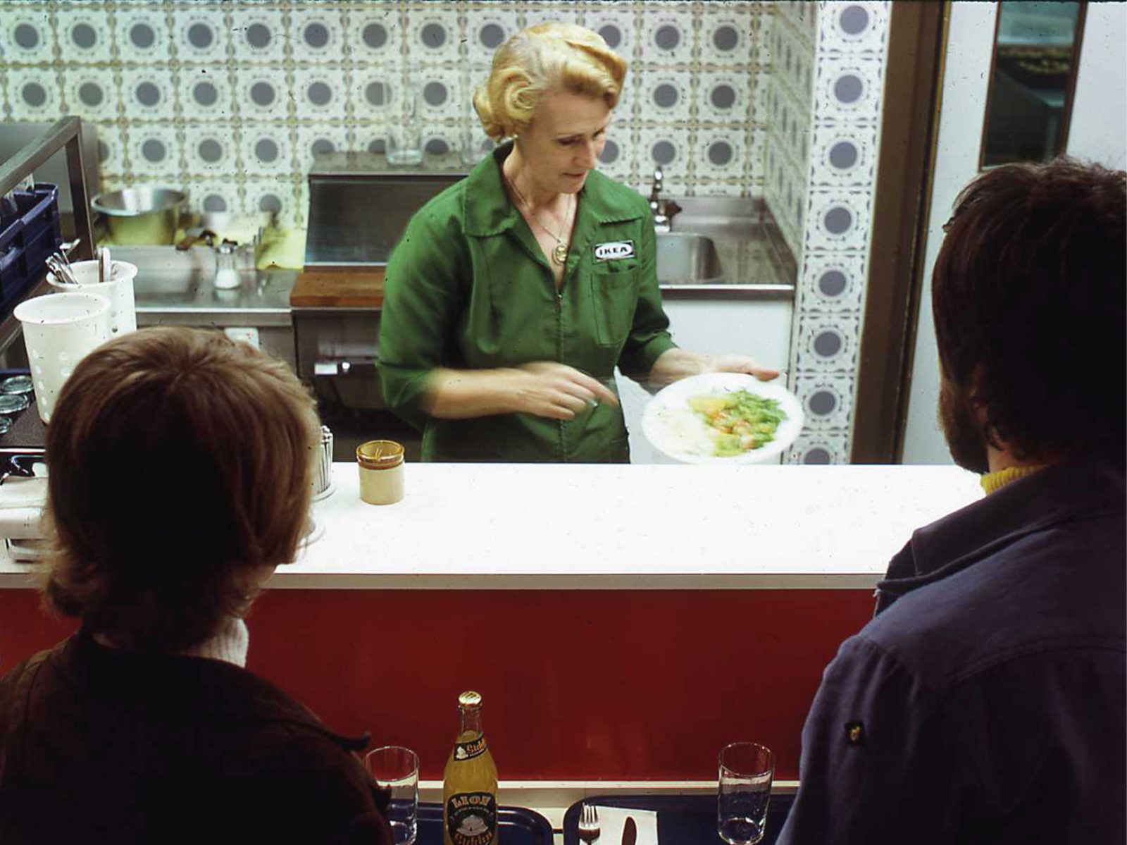 Un couple attend devant un comptoir self-service, assiette préparée par une femme blonde, uniforme vert IKEA années 1960.