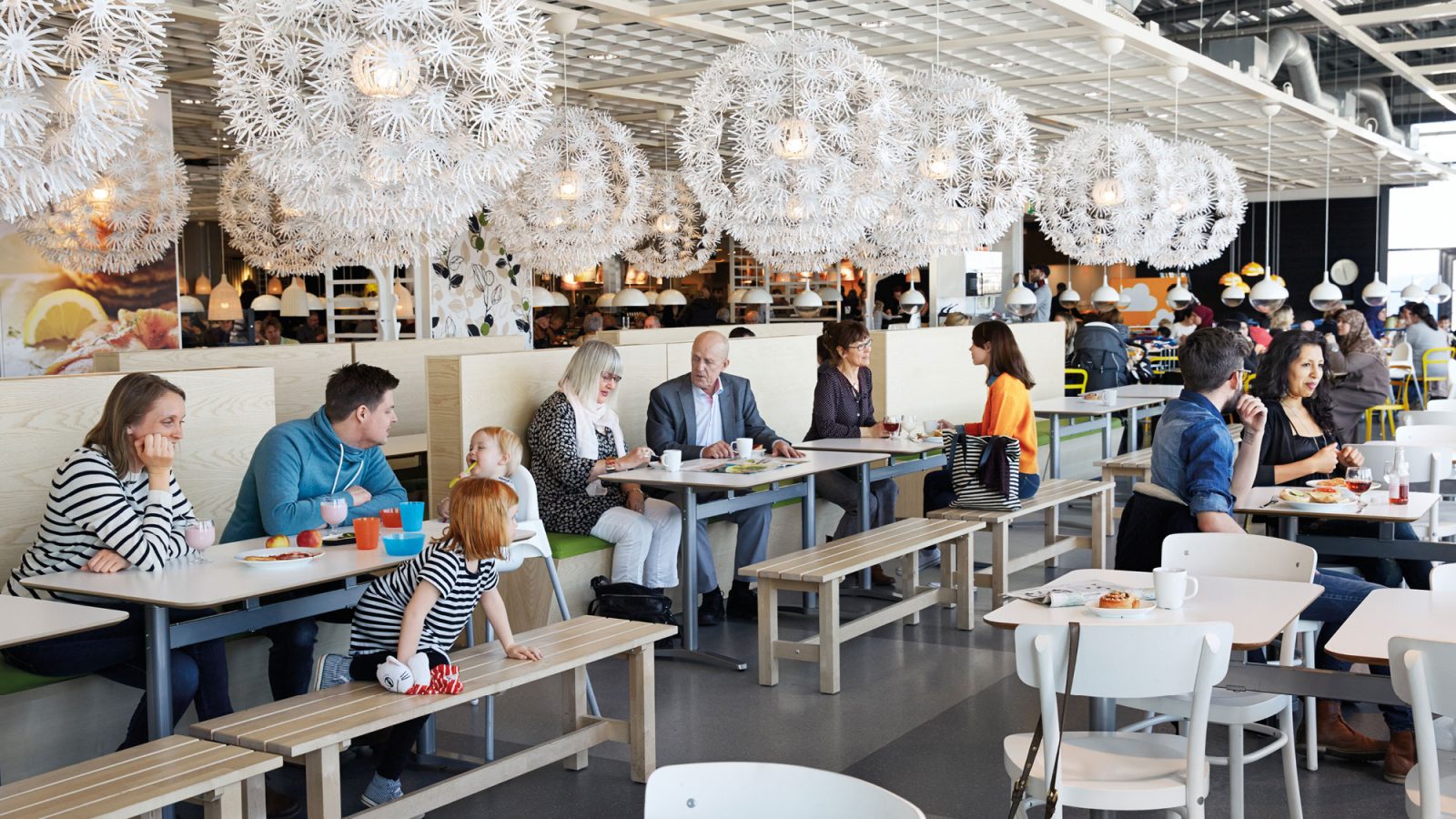 Families with children and couples eat under large white lamps in an airy contemporary IKEA restaurant with wooden furniture.