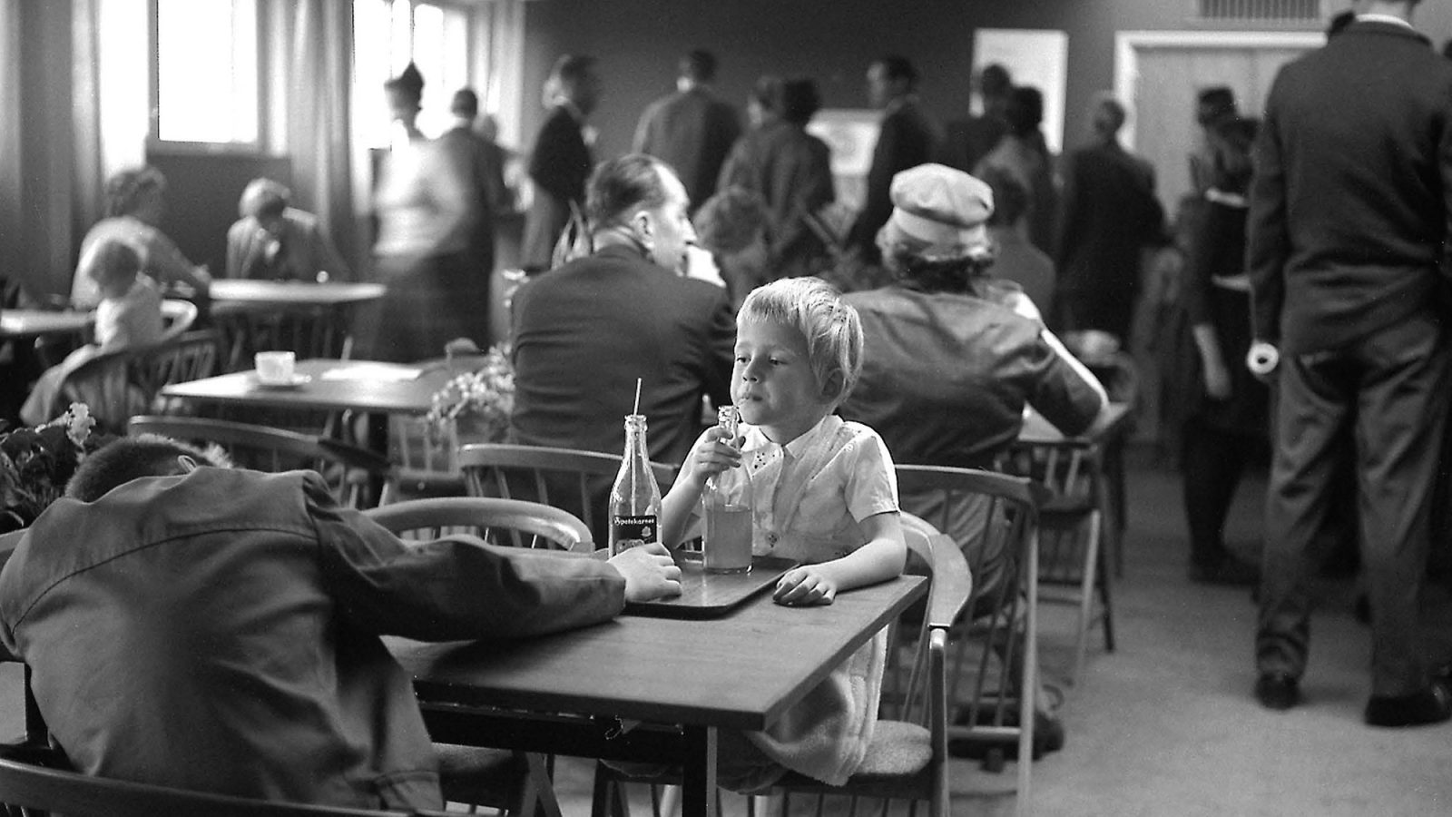 Child drinks soda from glass bottle with straw in a crowded restaurant with 1960s wood furnishings.