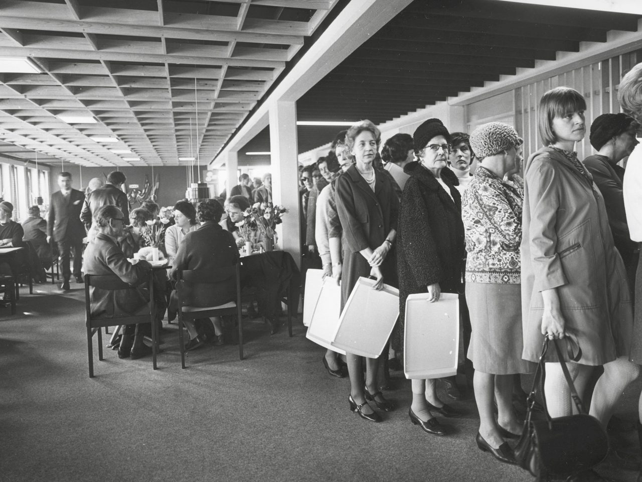 Women of different ages, dressed in 1960s style, stand in a restaurant queue holding trays.