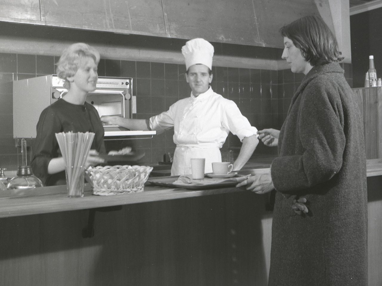 Woman in 1960s clothes stands at self-service counter, with serving staff including a chef standing by a radar oven.