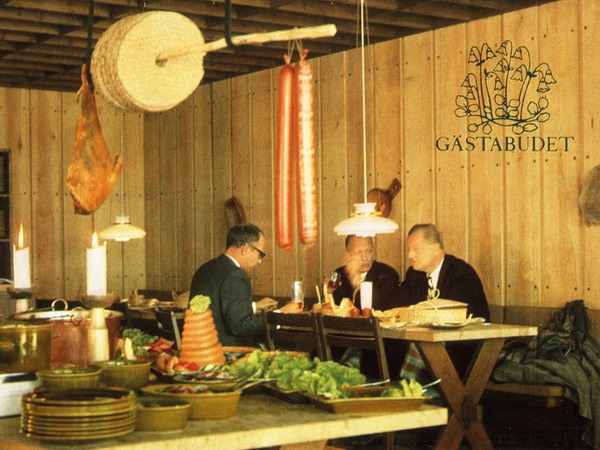 Three men in black suits sit in restaurant with wooden walls, in foreground round crispbread hangs on bar above a buffet table.