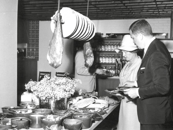 Homme en costume noir, femme avec chapeau et manteau style années 1960, remplissent leurs assiettes à un buffet.