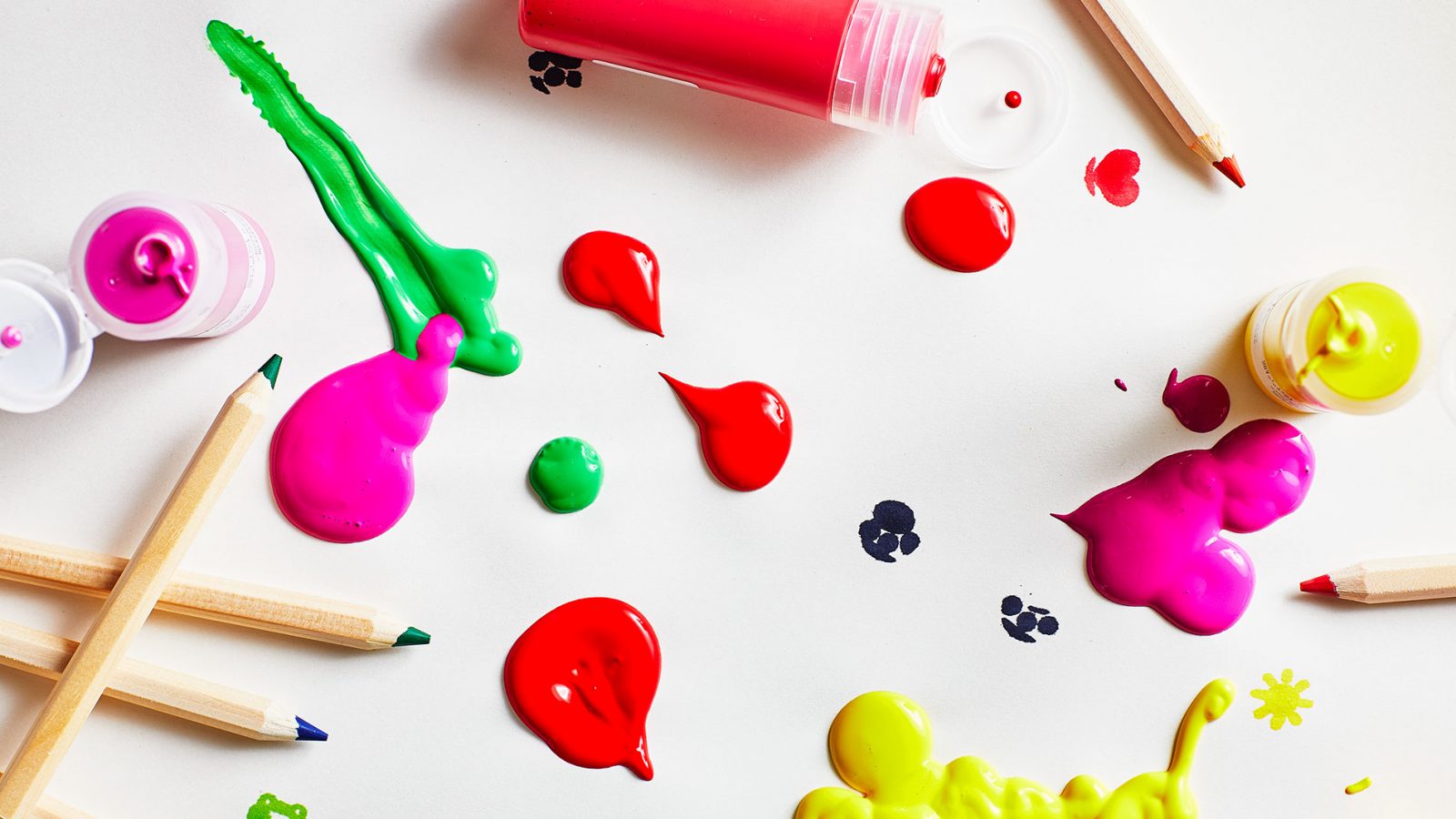 Wooden pencils in mixed colours and splashes of yellow, green, pink and red paint on a white surface.