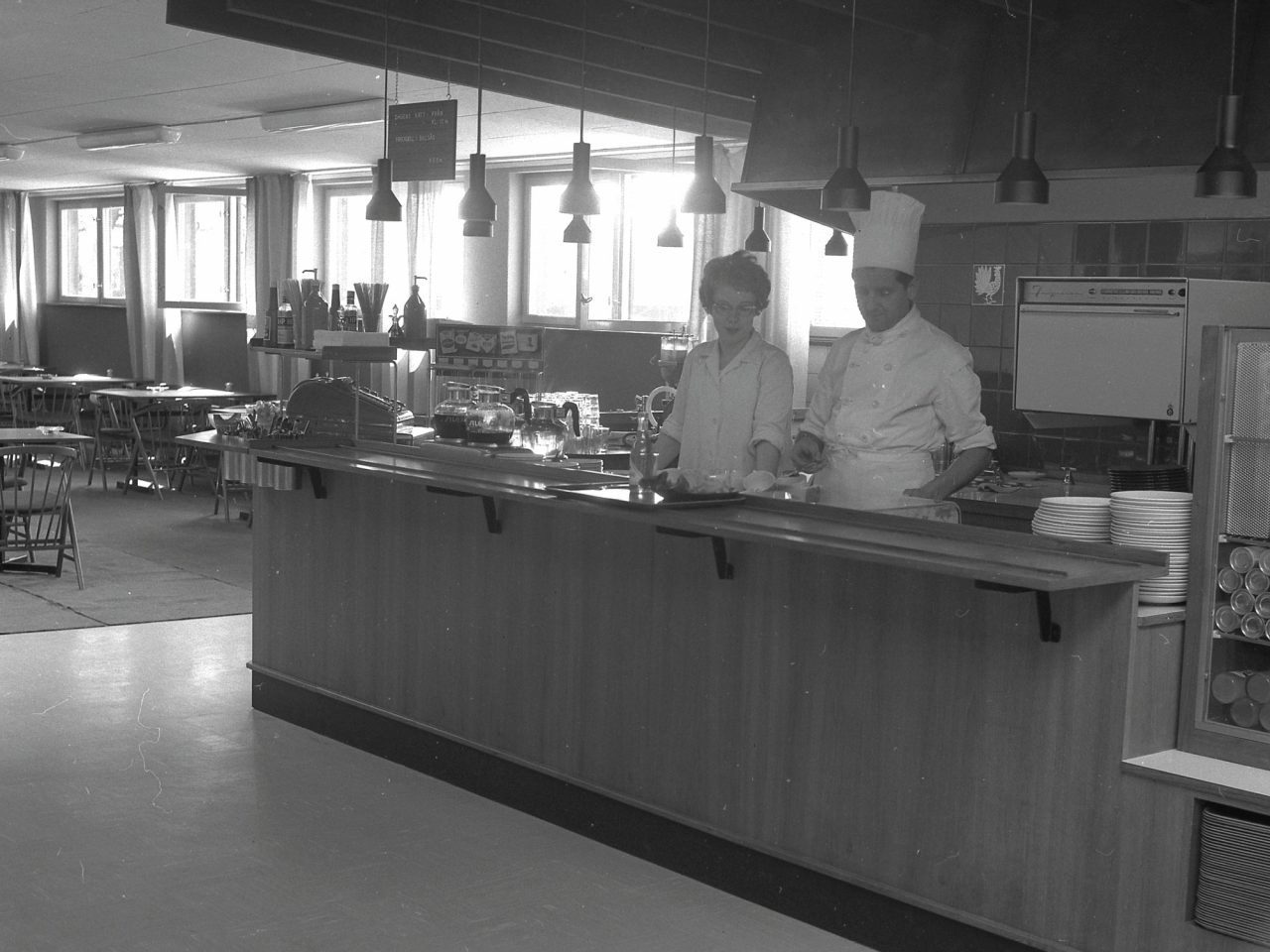 A chef and a female kitchen staff member stand in the self-service area of a restaurant with 1960s wood furnishings.