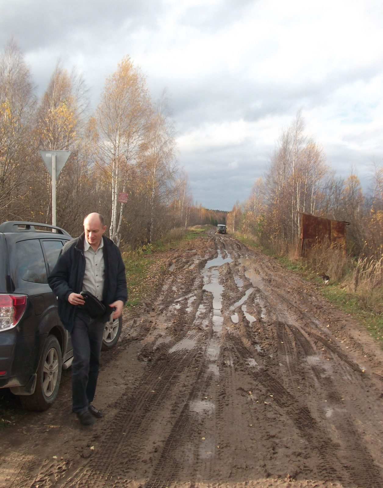 Man stands next to a car in a muddy dirt road, birch trees on each side.