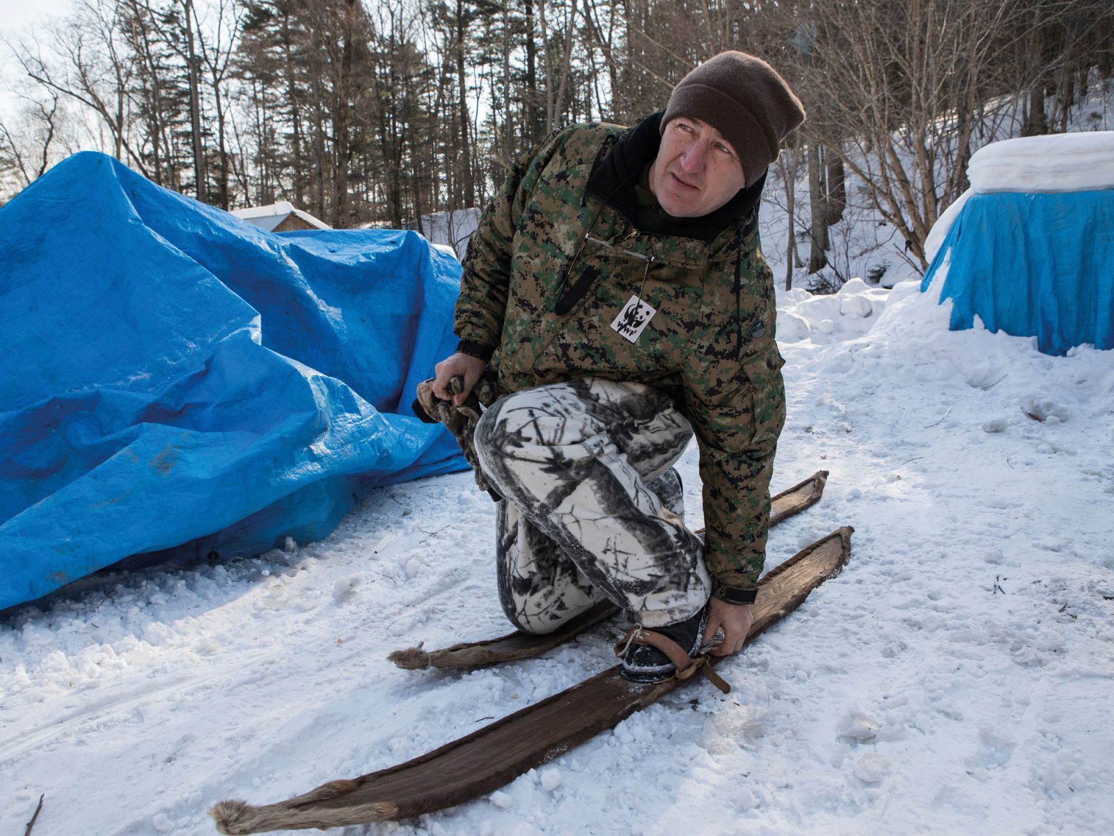 Man i varma kläder och mössa tar på sig gammaldags skidor på snötäckt mark, framför blåa tält.