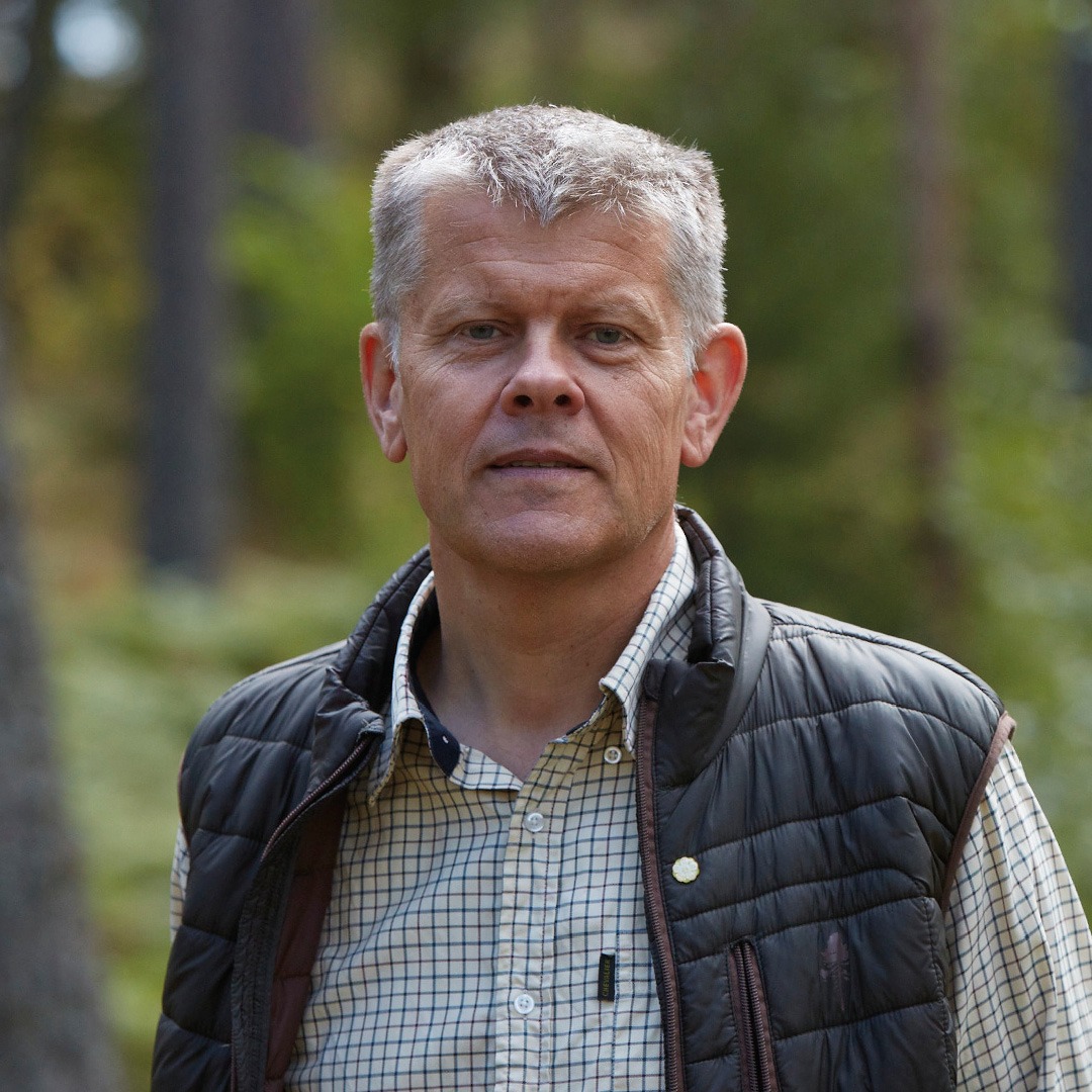 Grey-haired white man with checked shirt and a down vest stands in forest environment