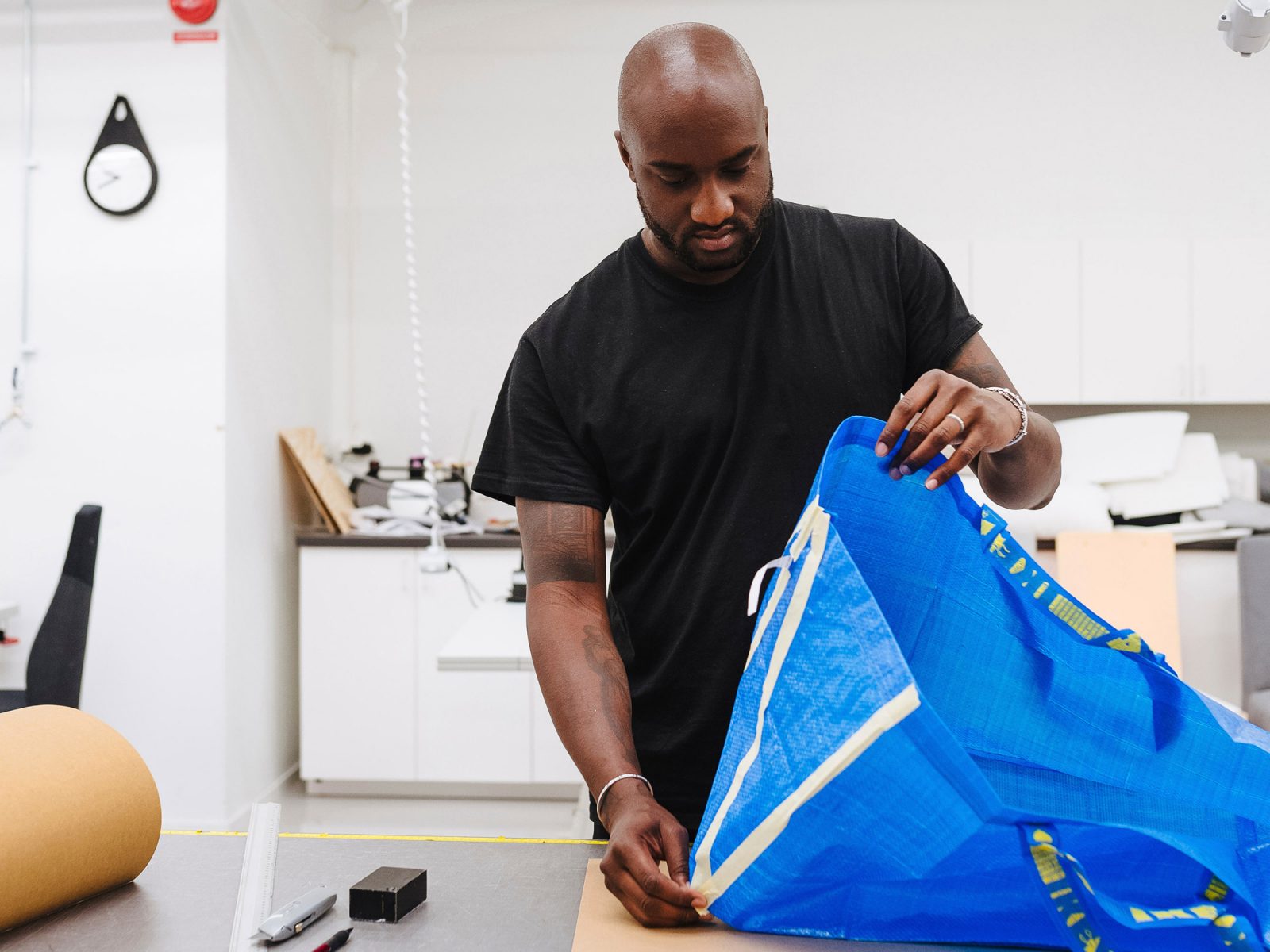 Un homme en T-shirt, tête rasée, Virgil Abloh, travaille à transformer un sac FRAKTA bleu dans un atelier.