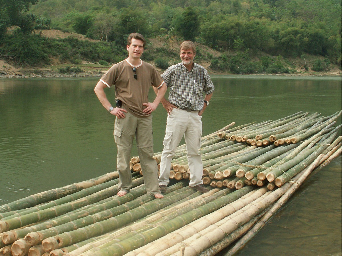 Two smiling men stand on a bamboo raft in a river, rainforest seen behind.