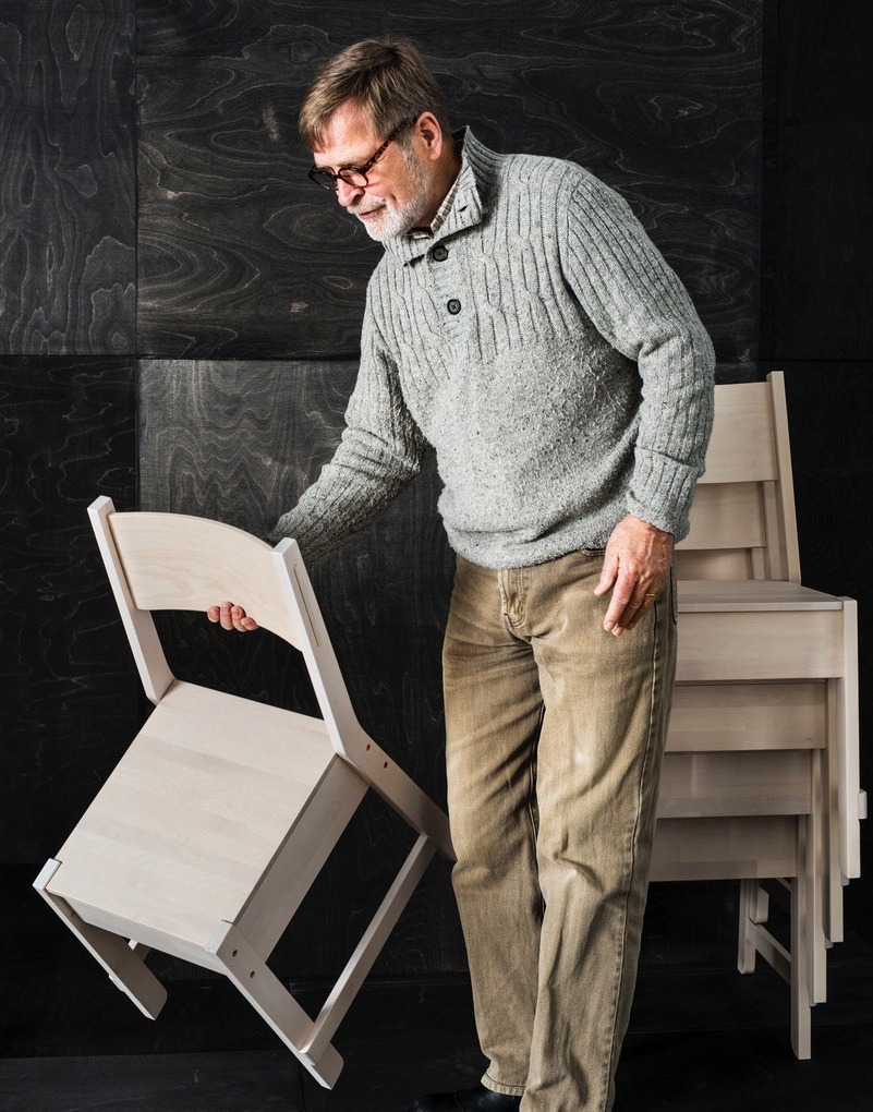 Grey-blond man in glasses and grey knit sweater lifts a chair made of wood.