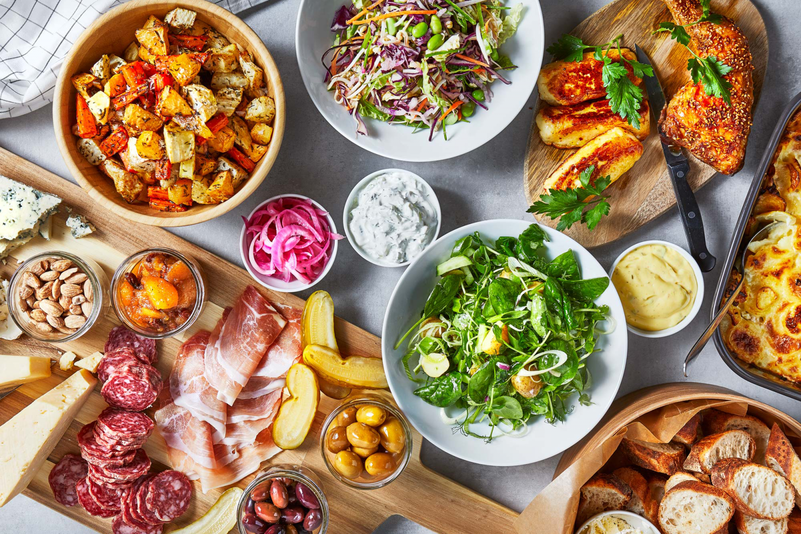 Table filled with salami, pickles, olives, sallad, potato sallad, bread, gravy and ham.