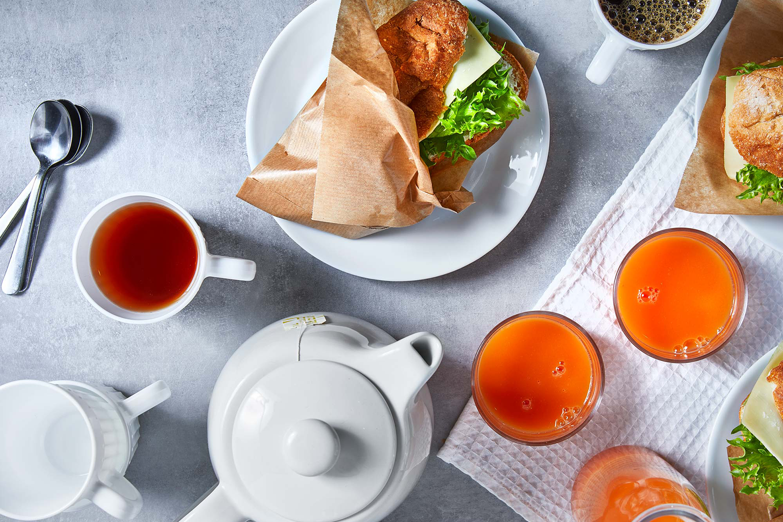 Tekanna, tekoppar, juiceglas fyllda med juice, ostmackor med sallad och kaffekoppar på ett dukat bord.