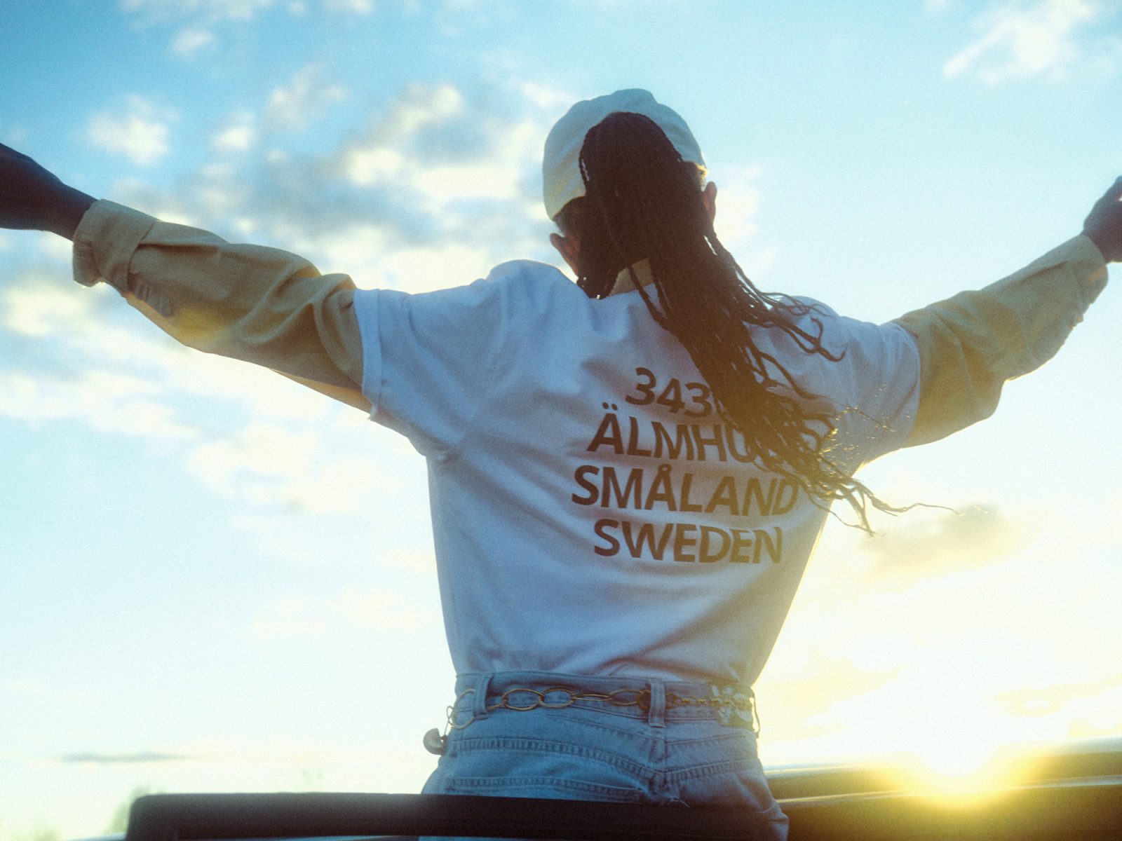 Kvinna som står i en bild och lutar sig emot bildörren. Hon sträcker händerna mot himmeln och är klädd i baggy jeans och en vit t-shirt med svart text: 343 36 ÄLMHULT SMÅLAND SWEDEN.