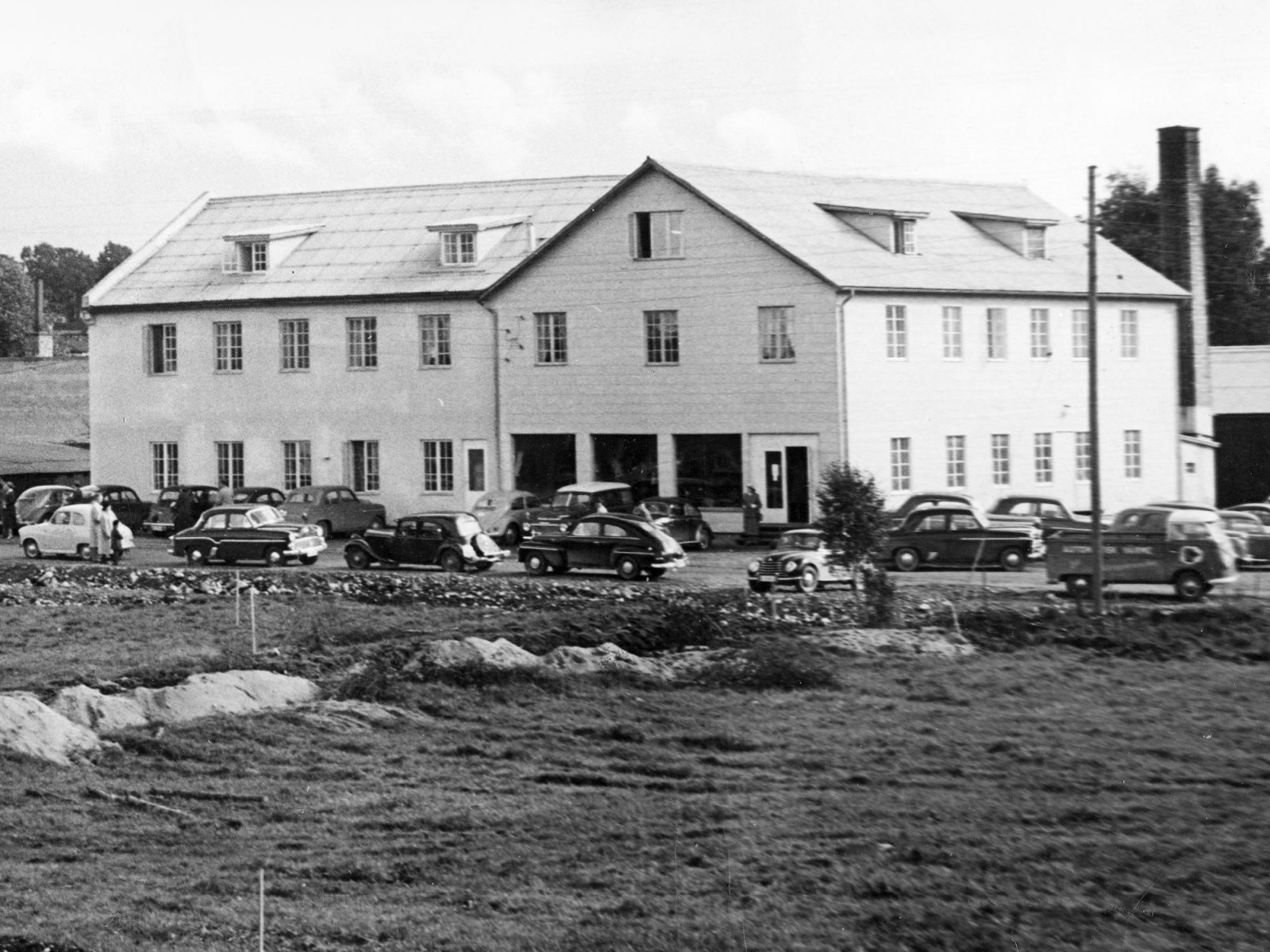 Large white two-storey building, 1950s cars parked outside, early-stage building site staked out in foreground.