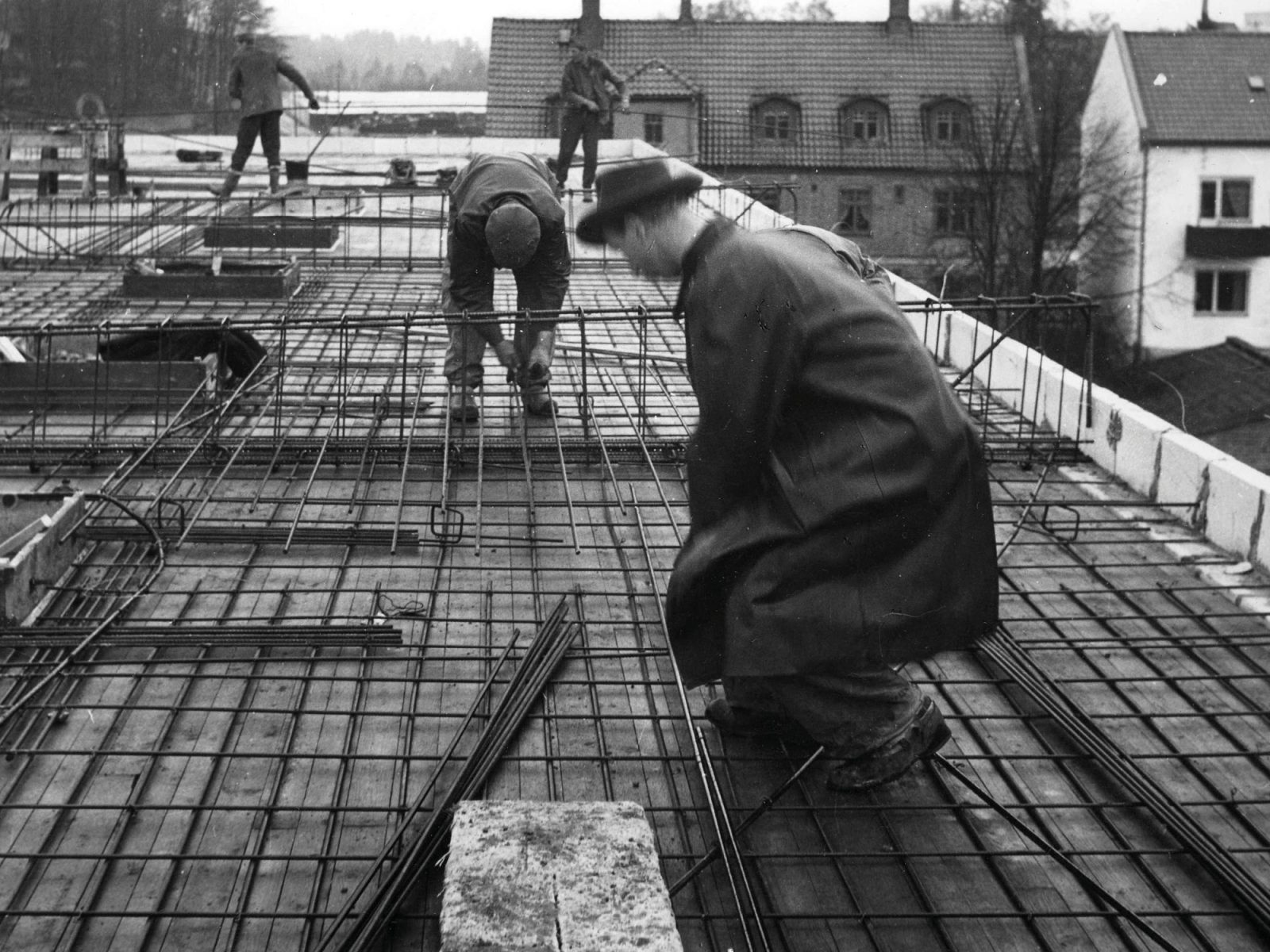 Mann im Stil der 1950er mit Hut und Mantel gemeinsam mit anderen auf einer Baustelle.